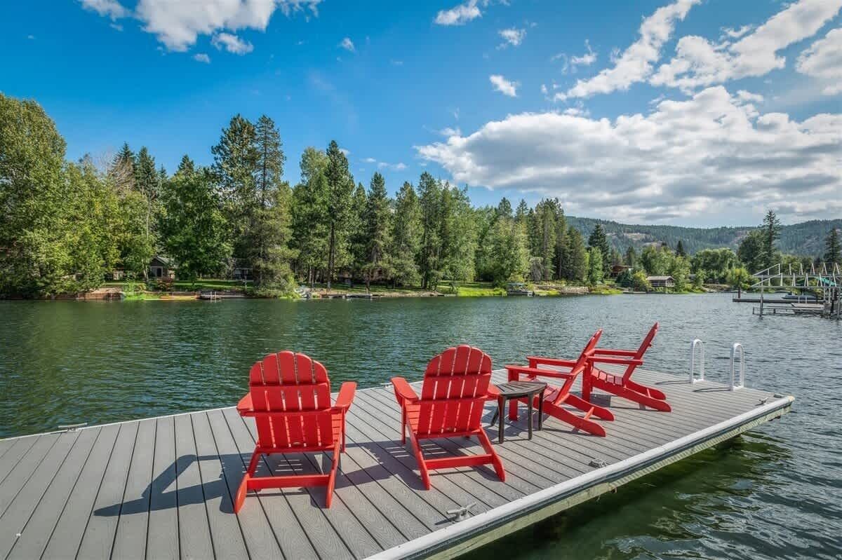 En träbrygga med röda Adirondack-stolar och ett litet bord, med utsikt över en sjö och en bakgrund av träd och kullar.