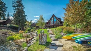 A lodge with a stone pathway leading to a wooden deck, surrounded by a natural landscape with a pond and kayaks. - Waterfront Log Home, Dock, Hot Tub, and Pickleball (Sagle)