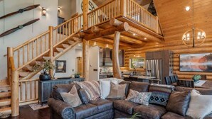 A spacious living area with a large wooden staircase, leather sofas, and a modern kitchen in the background. - Waterfront Log Home, Dock, Hot Tub, and Pickleball (Sagle)