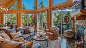 A log cabin interior with a fireplace, leather sofas, a coffee table, and large windows offering a view of the outdoors. - Waterfront Log Home, Dock, Hot Tub, and Pickleball (Sagle)