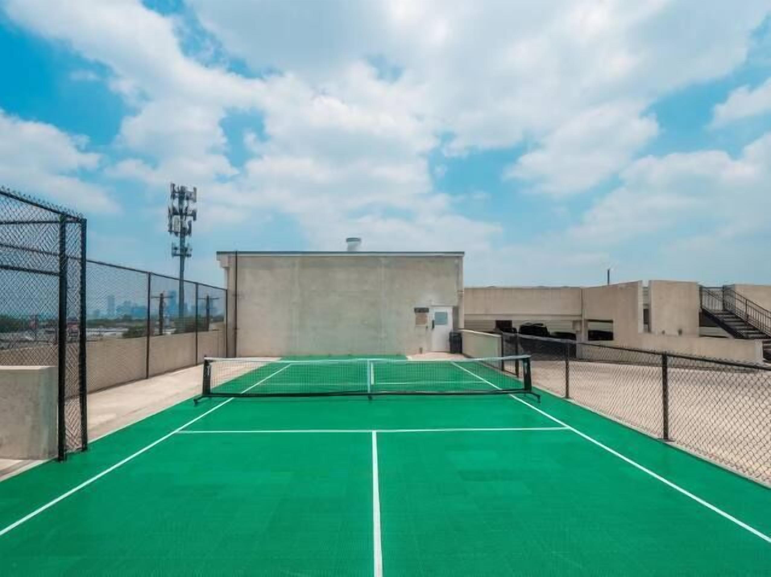 En udendørs tennisbane med net, omgivet af wirehegn, og en betonbygning i baggrunden.