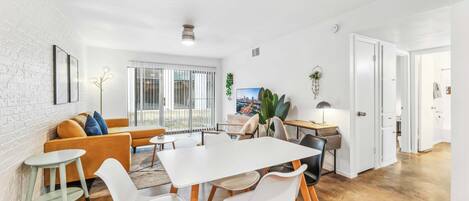 Una sala de estar moderna con una mesa y sillas blancas, un sofá y una mesa de centro. Hay un ventanal con persianas, una planta y un cuadro en la pared.