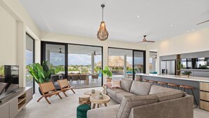 Amplia sala de estar con un sofá grande, sillas de madera y una mesa de comedor. Hay una cocina de diseño moderno y vistas al exterior.