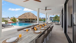 Zona de comedor junto a la piscina con una mesa larga dispuesta para el desayuno, con vistas a una piscina y una estructura con techo de paja.