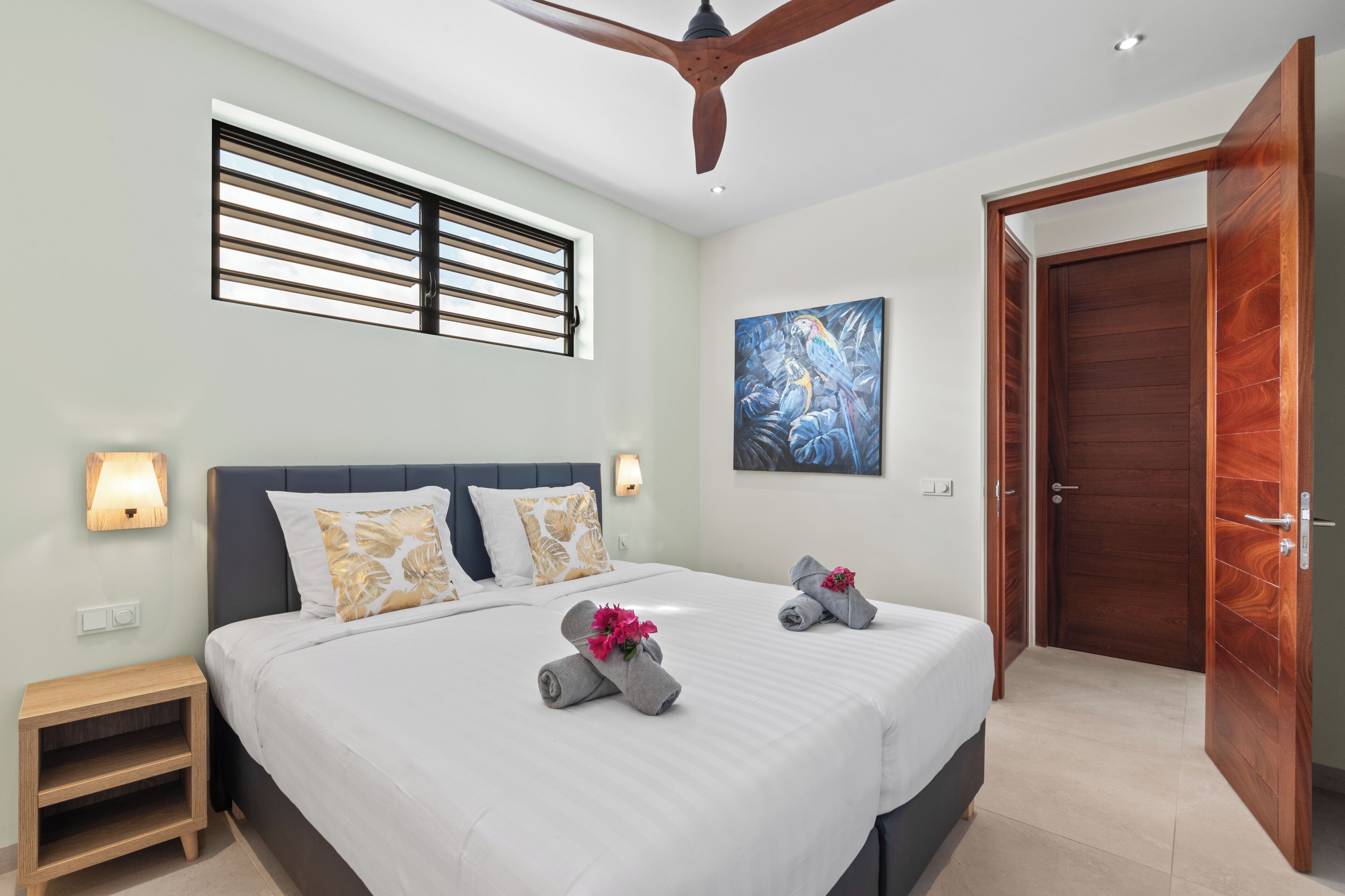 Une chambre d’hôtel moderne avec un grand lit, un ventilateur de plafond, une fenêtre avec des stores, un tableau au mur et une porte en bois.