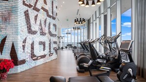 Une salle de sport équipée de tapis de course, de vélos d’appartement et d’appareils de musculation. Un mur de briques est décoré d’une fresque et de grandes fenêtres offrent une vue sur le ciel.