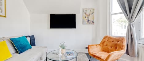 Una sala moderna con un sofá, una mesa de centro redonda de vidrio, una butaca naranja y un televisor de pantalla plana, además de un ventanal con cortinas.