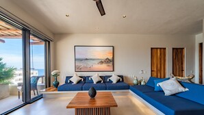 A modern living room with a large window, a blue sofa set, a wooden coffee table, and a painting of a beach scene. - Private Oceanview Splash Pool & Beach Access (Huatulco)