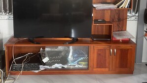 En stue med et stort fladskærms TV på et træ-tv-møbel, en reol med bøger og elektronisk udstyr, samt et vindue med gardiner.