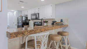 A kitchen with white cabinets, a granite countertop, a stainless steel refrigerator, and two bar stools.