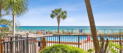 A poolside view with a clear view of the ocean, palm trees, and a fenced area with lounge chairs.