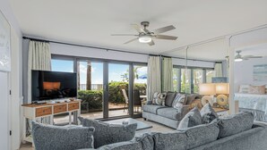 A modern living room with a flat-screen TV, grey sofas, a ceiling fan, and a sliding glass door leading to an outdoor area.