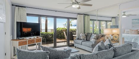 A modern living room with a flat-screen TV, grey sofas, a ceiling fan, and a sliding glass door leading to an outdoor area.