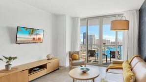 A modern living room with a TV, sofa, coffee table, and a balcony with a city view. - Best Views Balcony 2BR - Gym & Pool (Hallandale Beach)
