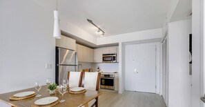 A modern kitchen with stainless steel appliances, a dining area with a wooden table set for four, and a white door leading to another room. - Stylish 2BR Apt w/Views (Pool & Gym) (Hallandale Beach)