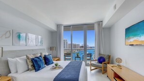 A modern hotel room with a large bed, a desk, a chair, and a view of a cityscape through a large window. - Stylish 2BR Apt w/Views (Pool & Gym) (Hallandale Beach)