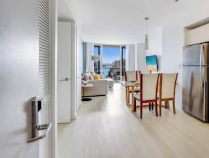 Una cocina moderna con un refrigerador de acero inoxidable, una zona de comedor con mesa y sillas, y una sala de estar con sofá y vistas a la ciudad.