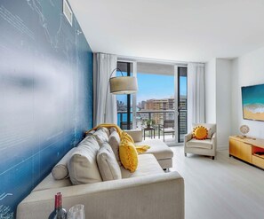 A modern living room with a sofa, armchair, and TV, a large window with a city view, and a wall with a blue design.