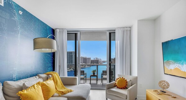 A modern living room with a sofa, armchair, and a view of a cityscape and water through large windows. - Stylish 2BR Apt w/Views (Pool & Gym) (Hallandale Beach)
