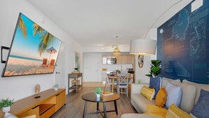 Un soggiorno moderno con una TV a schermo piatto, un tavolino rotondo e un divano con cuscini. C'è una zona pranzo con tavolo e sedie, e una cucina sullo sfondo.