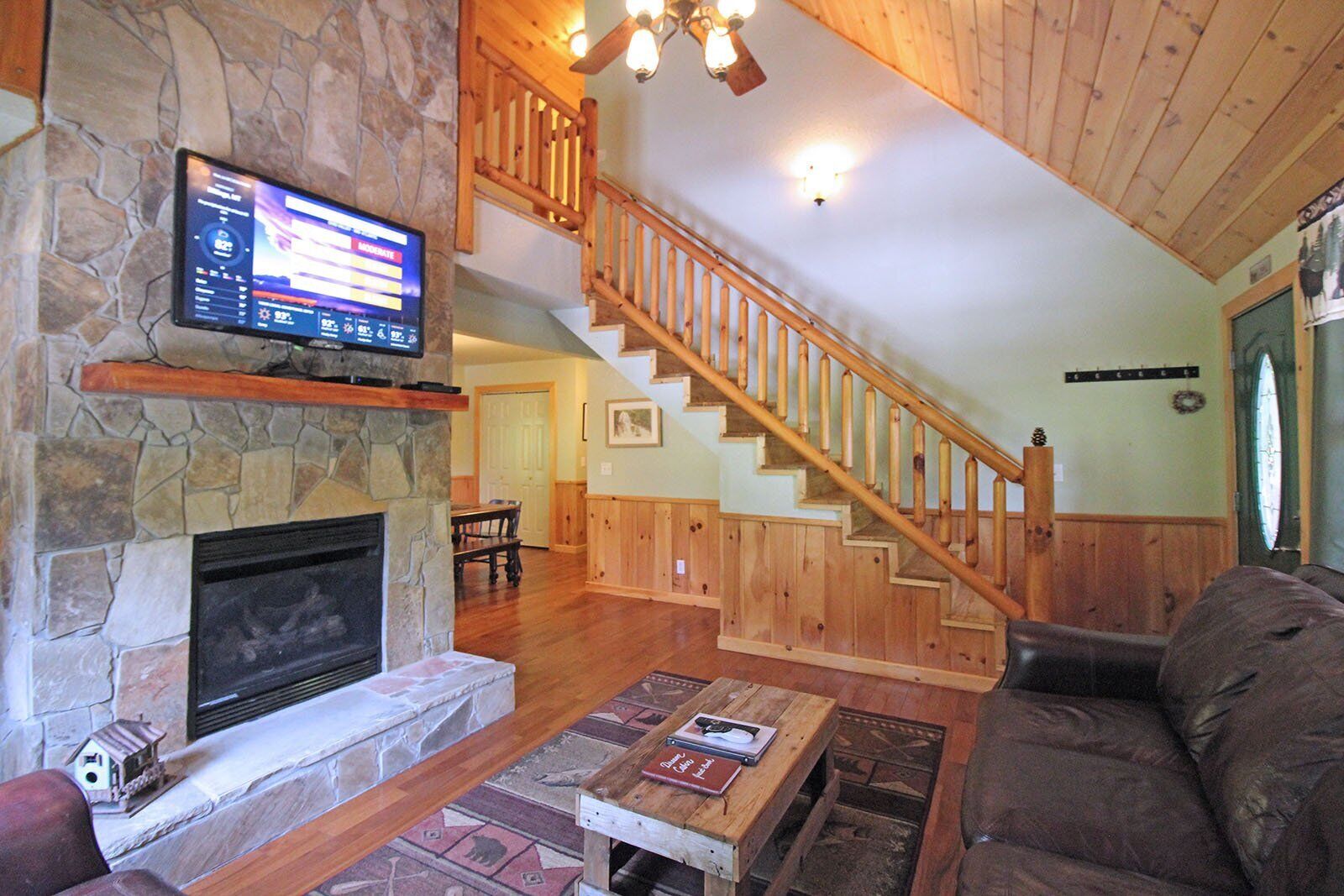 A cozy living room with a stone fireplace, wooden staircase, and a flat-screen TV mounted on the wall.