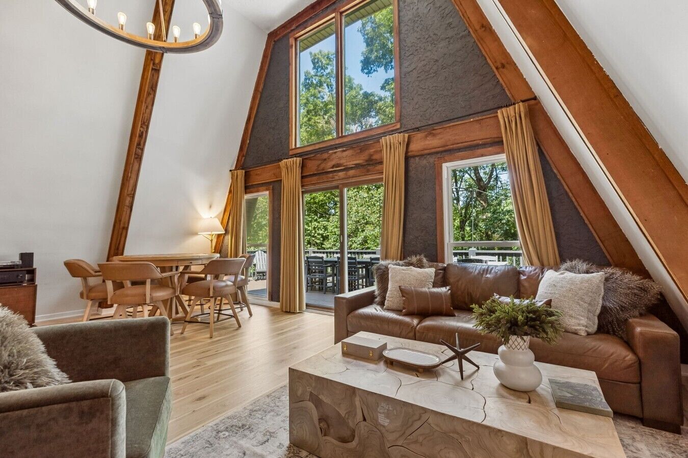 A spacious loft with a high ceiling, a dining area with wooden furniture, a leather sofa set, and large windows offering a view of the outdoors.