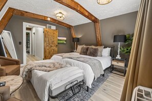 Un dormitorio con techo alto, una cama con ropa blanca, una puerta de madera y una mesita de noche con lámpara.
