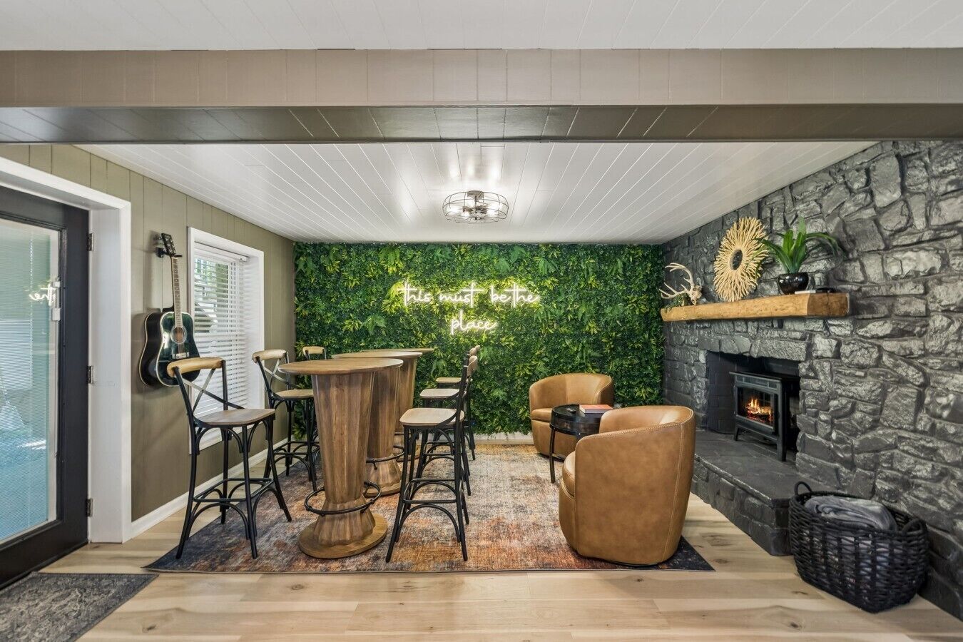 A cozy room with a stone fireplace, a green wall with a neon sign, and a wooden coffee table with chairs.