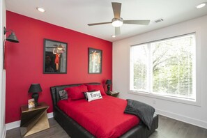A bedroom with a red wall, a bed with a red bedspread, a ceiling fan, and a large window.