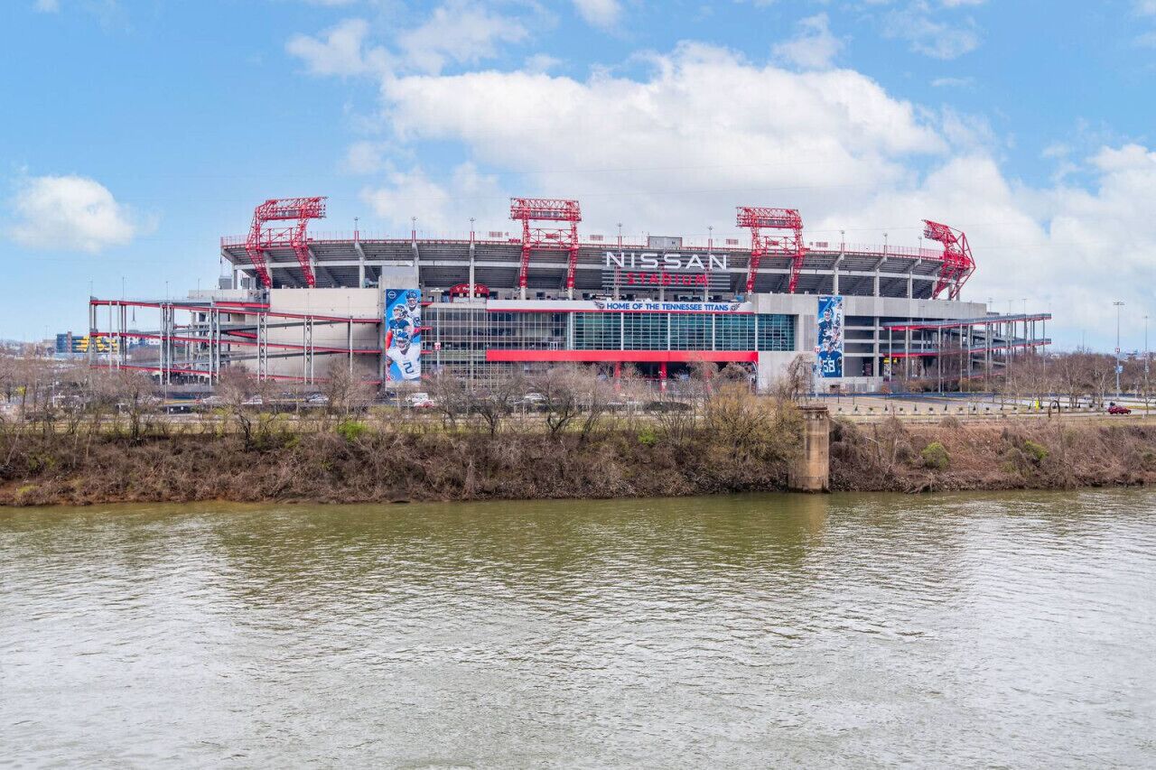 Un estadio en construcción con un gran letrero 'NISSAN', un río al frente y un cielo despejado.