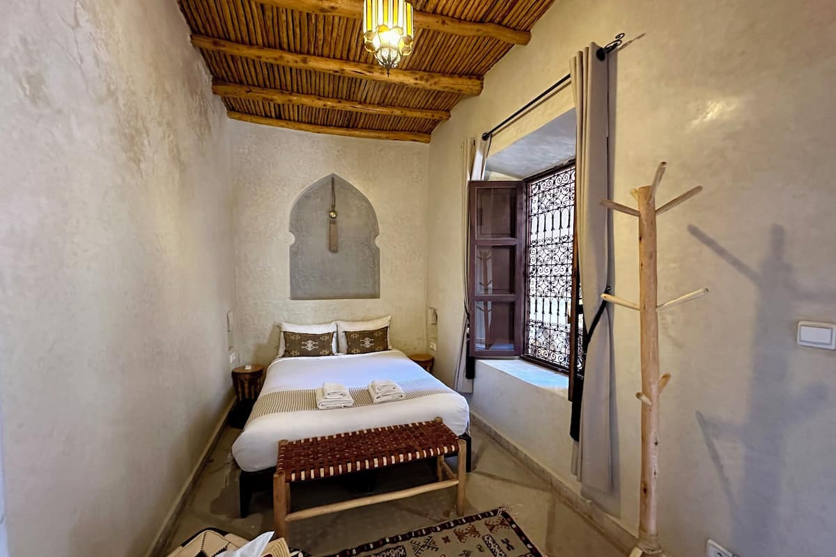 A bedroom with a wooden ceiling, a bed with white linens, a bench, a patterned rug, and a window with decorative bars.