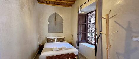 A bedroom with a wooden ceiling, a bed with white linens, a bench, a patterned rug, and a window with decorative bars.