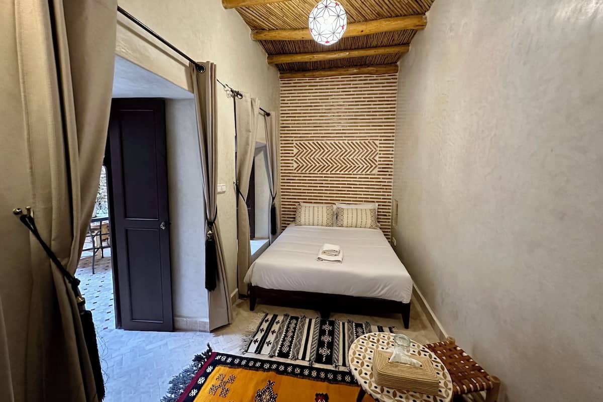 A narrow room with a bed, a patterned wall, a woven ceiling, and a door leading outside.