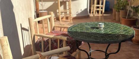 A wooden chair with a patterned cushion, a round table with a green mosaic top, and a potted plant in a sunlit outdoor area with a tiled floor and a wooden pergola in the background.