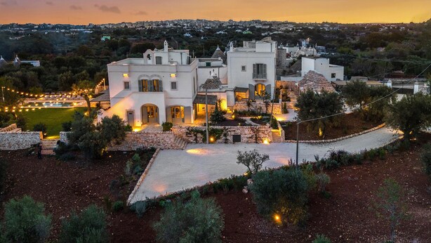 A large, white building with multiple sections, surrounded by landscaped gardens and a view of a cityscape in the background. - 3643 Masseria Stella del Monte by Perle di Puglia (Cisternino)