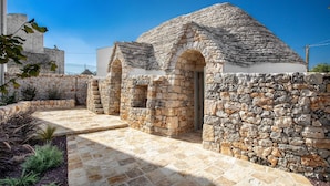 A stone building with a sloped roof and arched doorways, surrounded by a stone wall and a paved pathway. - 3643 Masseria Stella del Monte by Perle di Puglia (Cisternino)