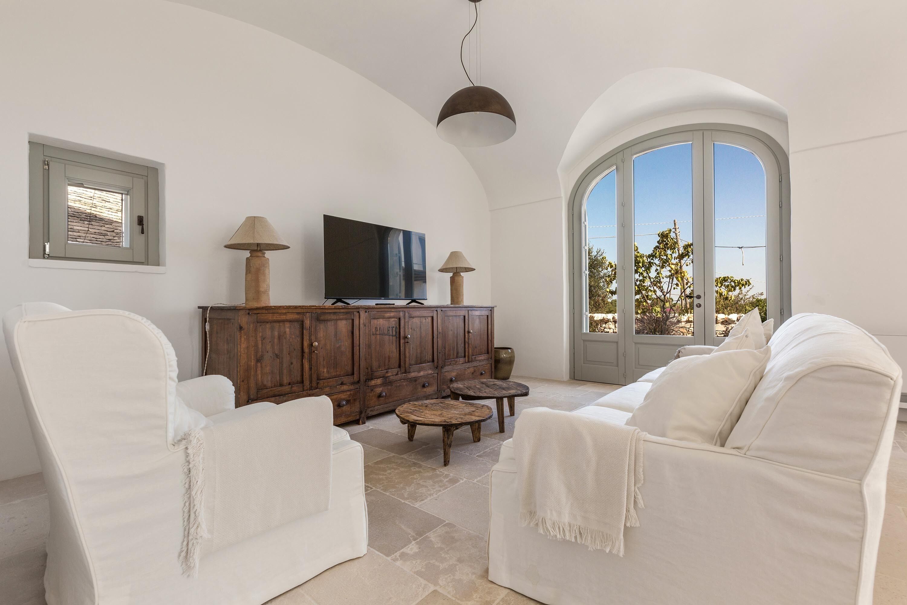 A modern living room with white furniture, a wooden cabinet, a flat-screen TV, and a large arched window offering a view of the outdoors.