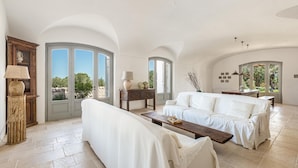 A spacious living room with a white sofa, a wooden coffee table, and a lamp. There are large windows with a view of greenery outside. - 3643 Masseria Stella del Monte by Perle di Puglia (Cisternino)