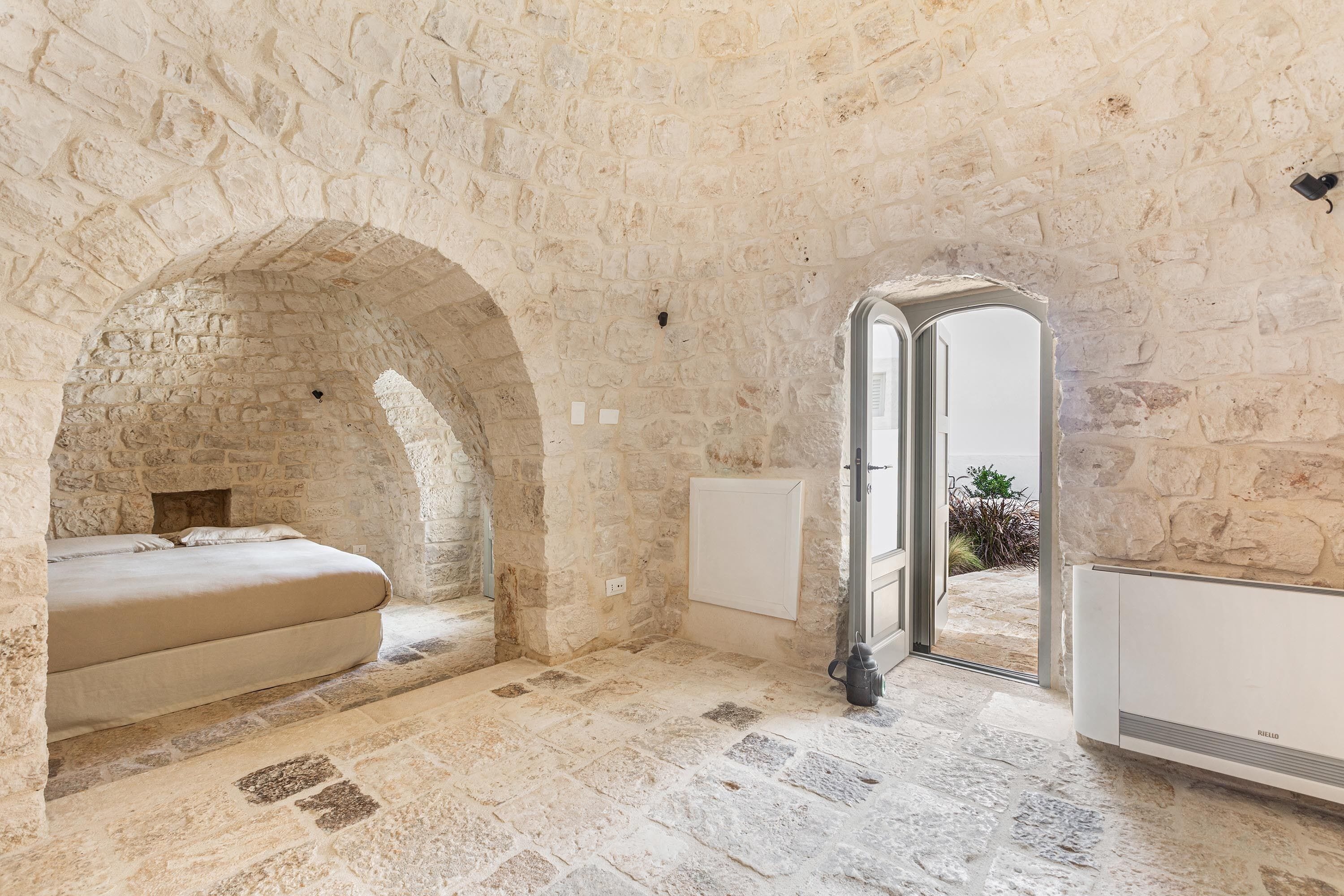 Quarto com paredes de pedra, uma cama, um radiador e uma porta aberta para o exterior.