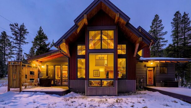 Una caba ña de troncos moderna con ventanas amplias, un porche cubierto y suelo cubierto de nieve.