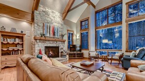 A cozy living room with a stone fireplace, leather sofas, and large windows offering a view of the outdoors.