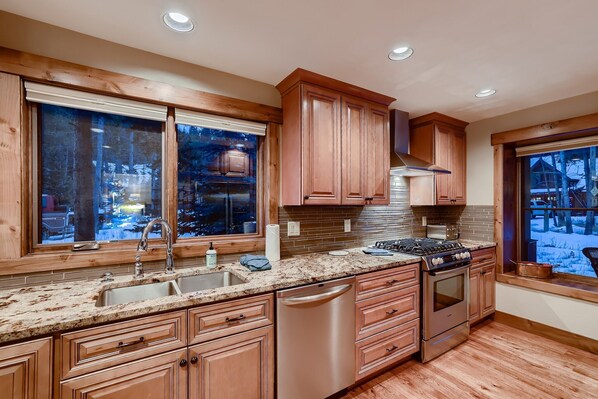A modern kitchen with wooden cabinets, a stainless steel dishwasher, and a large window with a view of a snowy landscape.