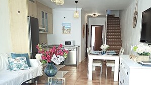 A spacious living room with a sofa, a coffee table with flowers, a dining area with a table and chairs, and a kitchen in the background.