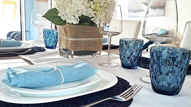 A dining table set with plates, cutlery, and blue glassware, with a basket of white flowers and a view of the sea through a window.