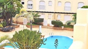 A pool area with a clear blue pool, surrounded by palm trees and cacti, with a building in the background.