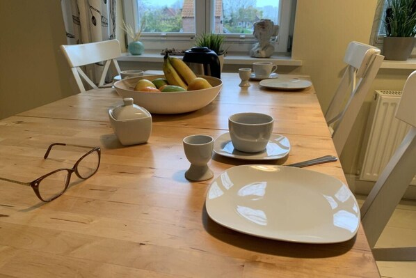 A dining table with a bowl of fruit, white plates, cups, and a teapot. - Ferienhaus Kleine Freiheit auf Fehmarn (Fehmarn OT Kopendorf)