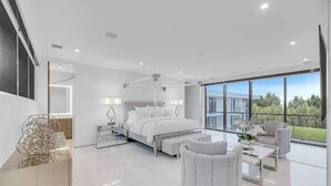 A modern bedroom with a large bed, a sitting area with a coffee table, and a view of a building and greenery outside. - Newly developed contemporary home situated on 1.5 acres. (Water Mill)