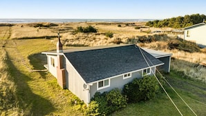 A single-story house with a sloped roof, a chimney, and a small attached garage. The property is surrounded by a grassy field with a view of the ocean in the background. - Hot Tub, Amazing Views, Ocean Front, Pets OK (Serenity) (Ocean Park)