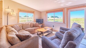 A spacious living room with a large L-shaped sofa, a coffee table, and two armchairs. There are two windows with blinds, a flat-screen TV, and a lamp on a stand. - Hot Tub, Amazing Views, Ocean Front, Pets OK (Serenity) (Ocean Park)