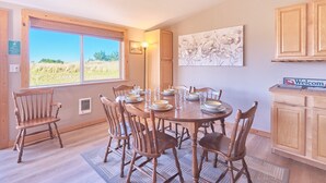 A dining area with a round table set for six, surrounded by wooden chairs. A window with a view of the outdoors is visible, and a 'Welcome' sign is on the wall. - Hot Tub, Amazing Views, Ocean Front, Pets OK (Serenity) (Ocean Park)